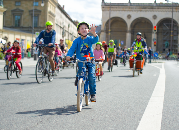 Kidical Mass Titelbild Kinder fahren mit dem Fahrrad bei einer Familien-Fahrraddemo. Ein Kind im Vordergrund winkt in die Kamera.